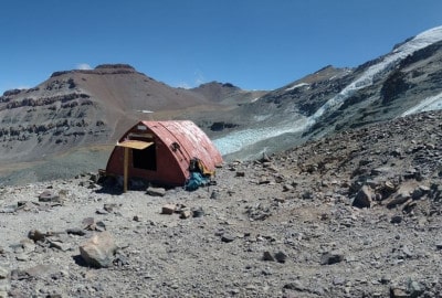 Una pequeña cabaña en la cima de una montaña con el Cerro El Plomo al fondo.